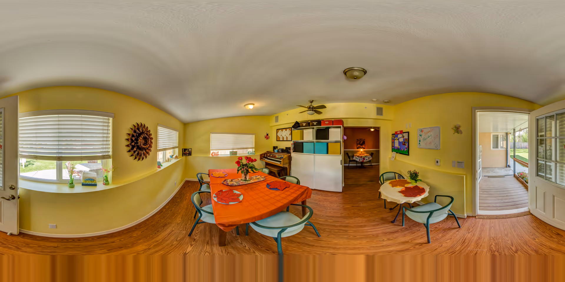 A bright, cozy common area in an assisted living facility with yellow walls and wood flooring. There is a rectangular table covered with an orange tablecloth and set with plates and napkins, surrounded by green chairs. A smaller round table with a white tablecloth and a plant is nearby. Windows with blinds let in natural light, and a door opens to an outdoor patio. The room features a piano, colorful storage cubes, a ceiling fan, and various wall decorations including a bulletin board and artwork.
