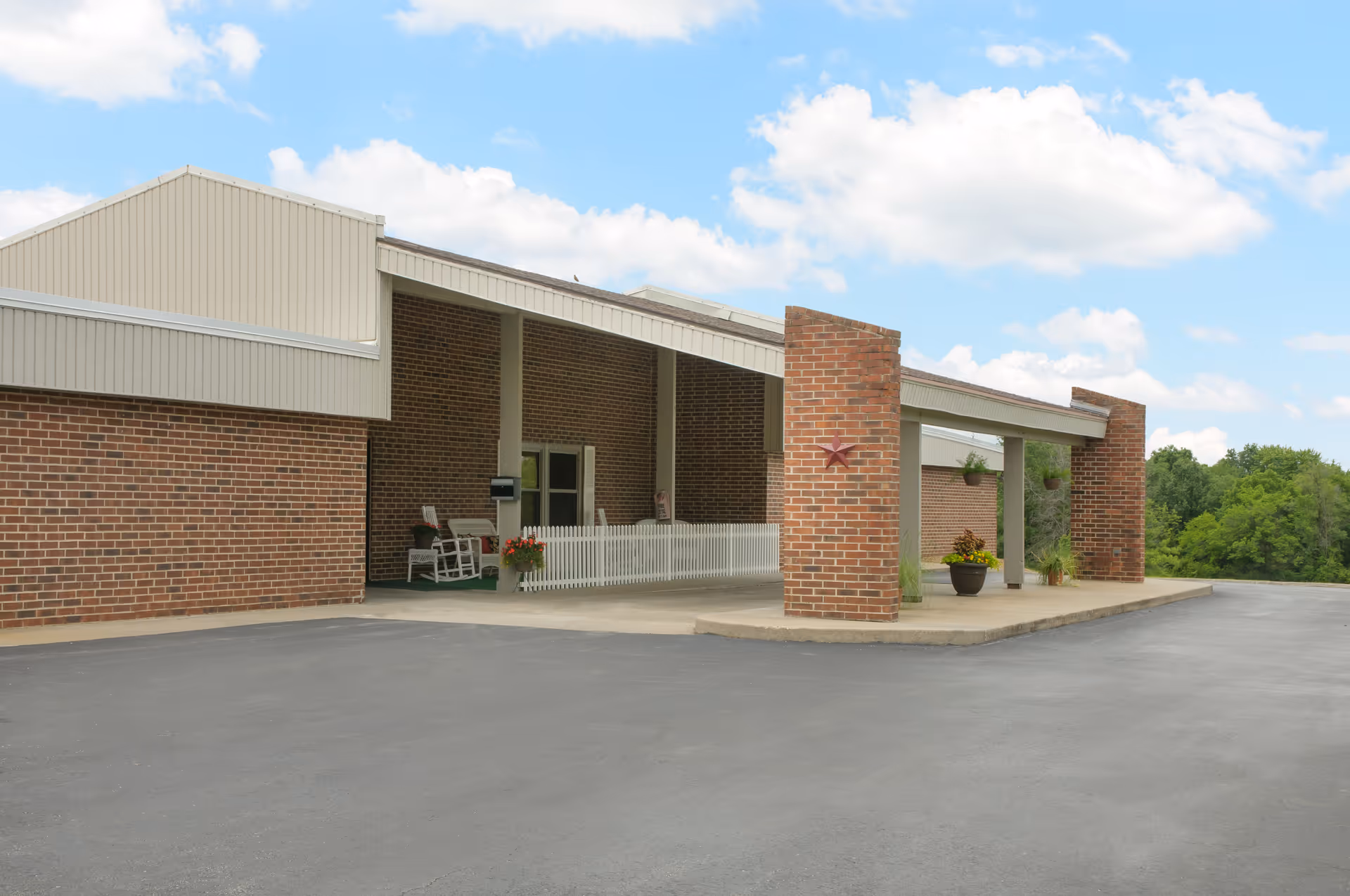 Covered entrance and porte-cochere of a brick building with potted plants and rocking chairs under a blue sky.