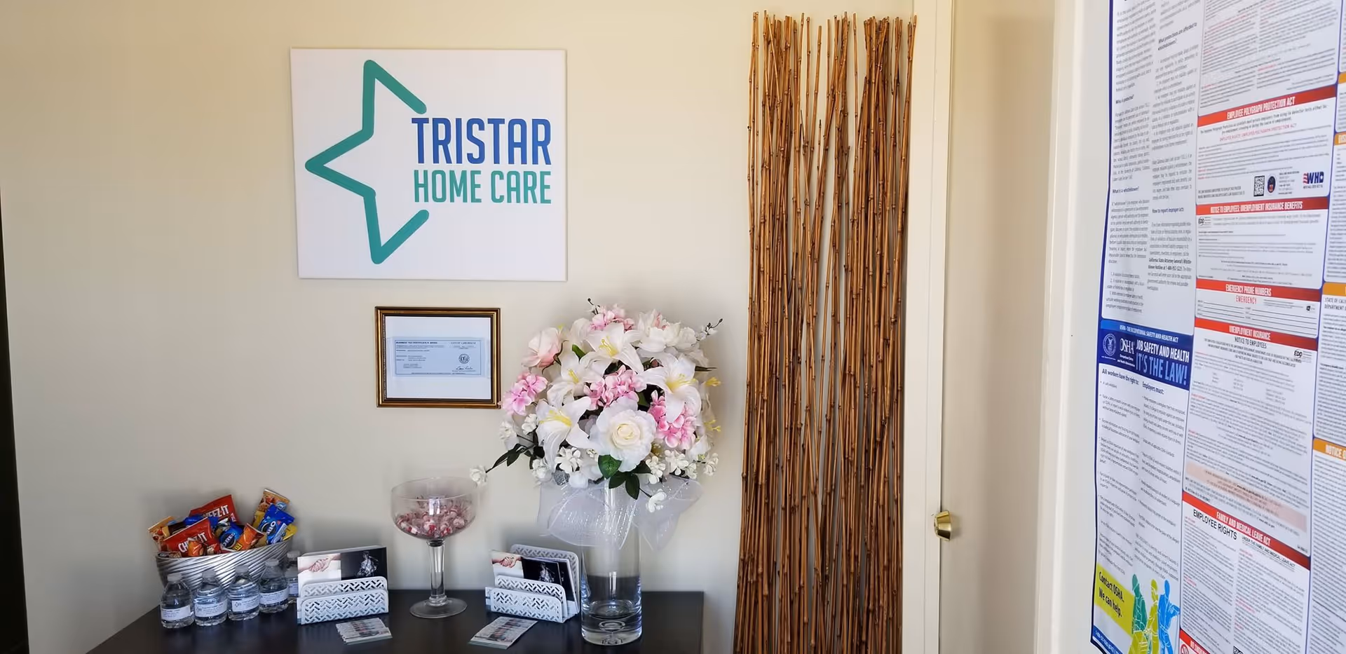 Reception area with Tristar Home Care sign, flowers, bamboo divider, snacks, and posters.