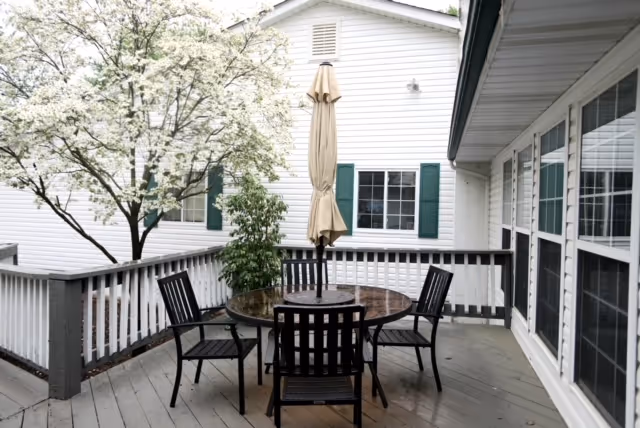 Outdoor deck with round table, chairs, and closed umbrella beside a white building.