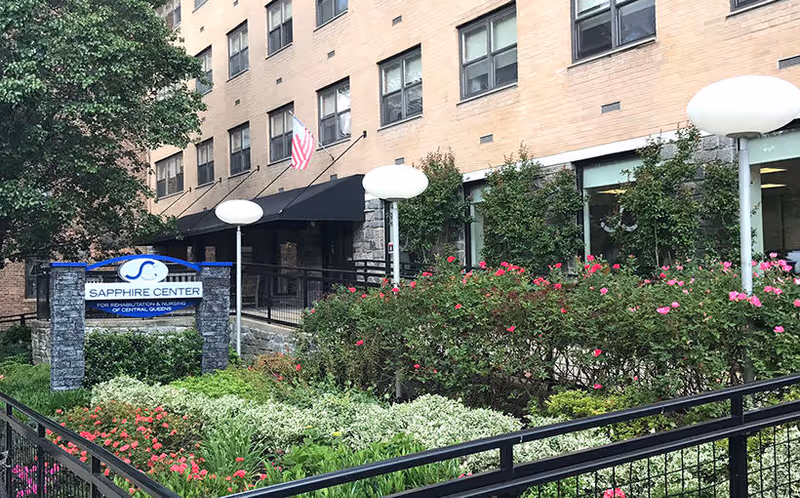 Front entrance of the Sapphire Center for Rehabilitation and Nursing showing the facility sign, landscaped garden, American flag, and multi-story brick building.