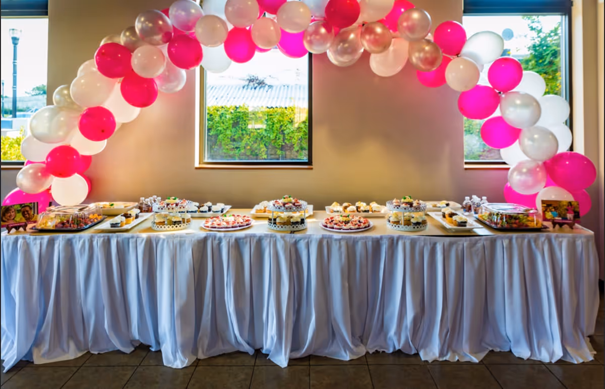 Dessert table with cakes and pastries beneath pink and white balloons in an interior room.