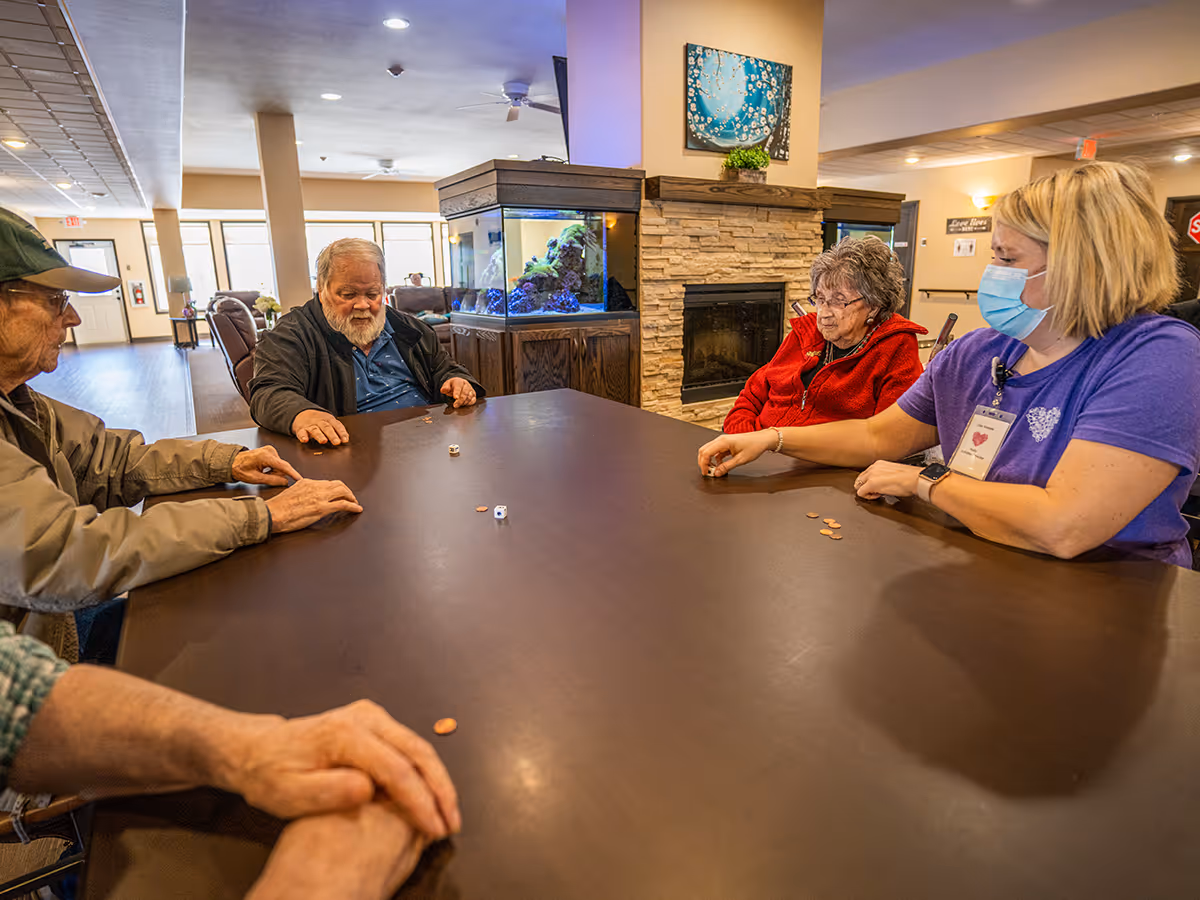 Seniors and a masked staff member sit around a large table playing a dice-and-penny game in a communal living room with an aquarium and fireplace.