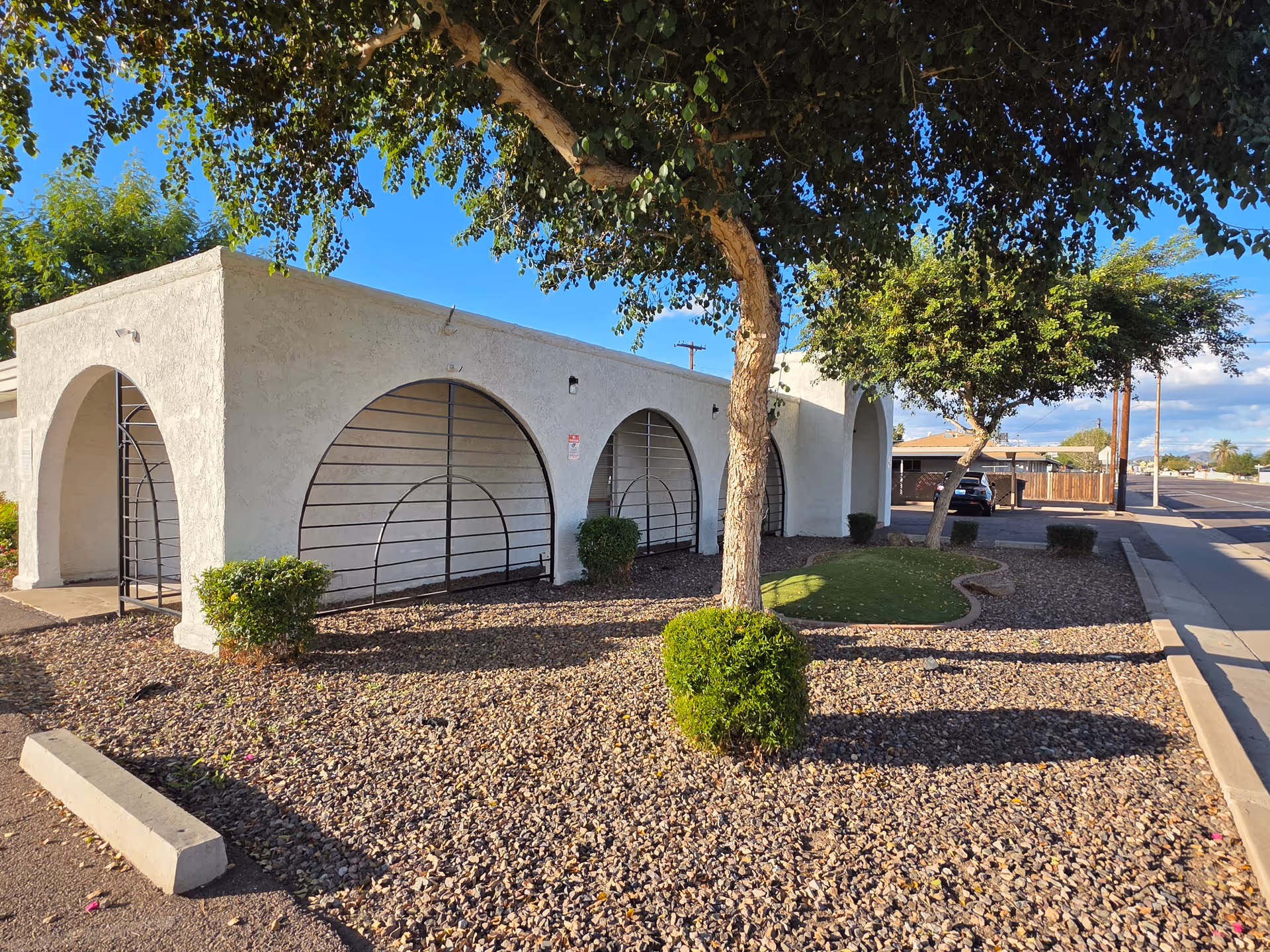 White stucco building with arched entrances, desert landscaping, and trees along a sunny street.