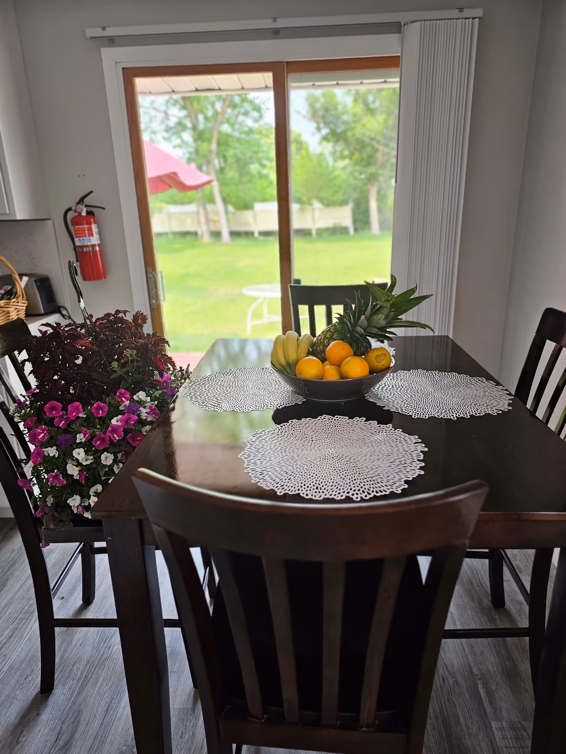 Wooden dining table with fruit bowl and crocheted placemats, facing sliding glass doors.