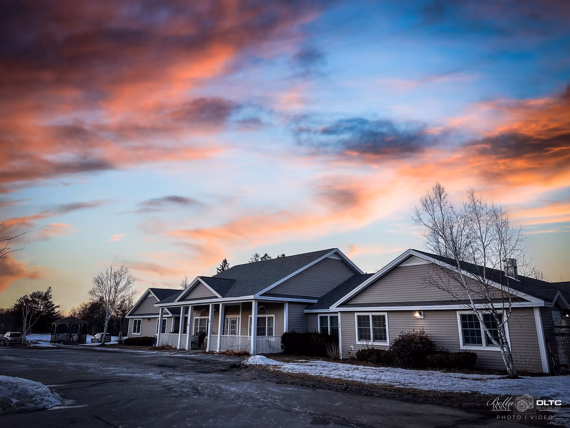 Exterior view of a single-story senior living facility building with a porch and multiple windows, set against a colorful sunset sky with pink and blue clouds. There is some snow on the ground and bare trees around the building.
