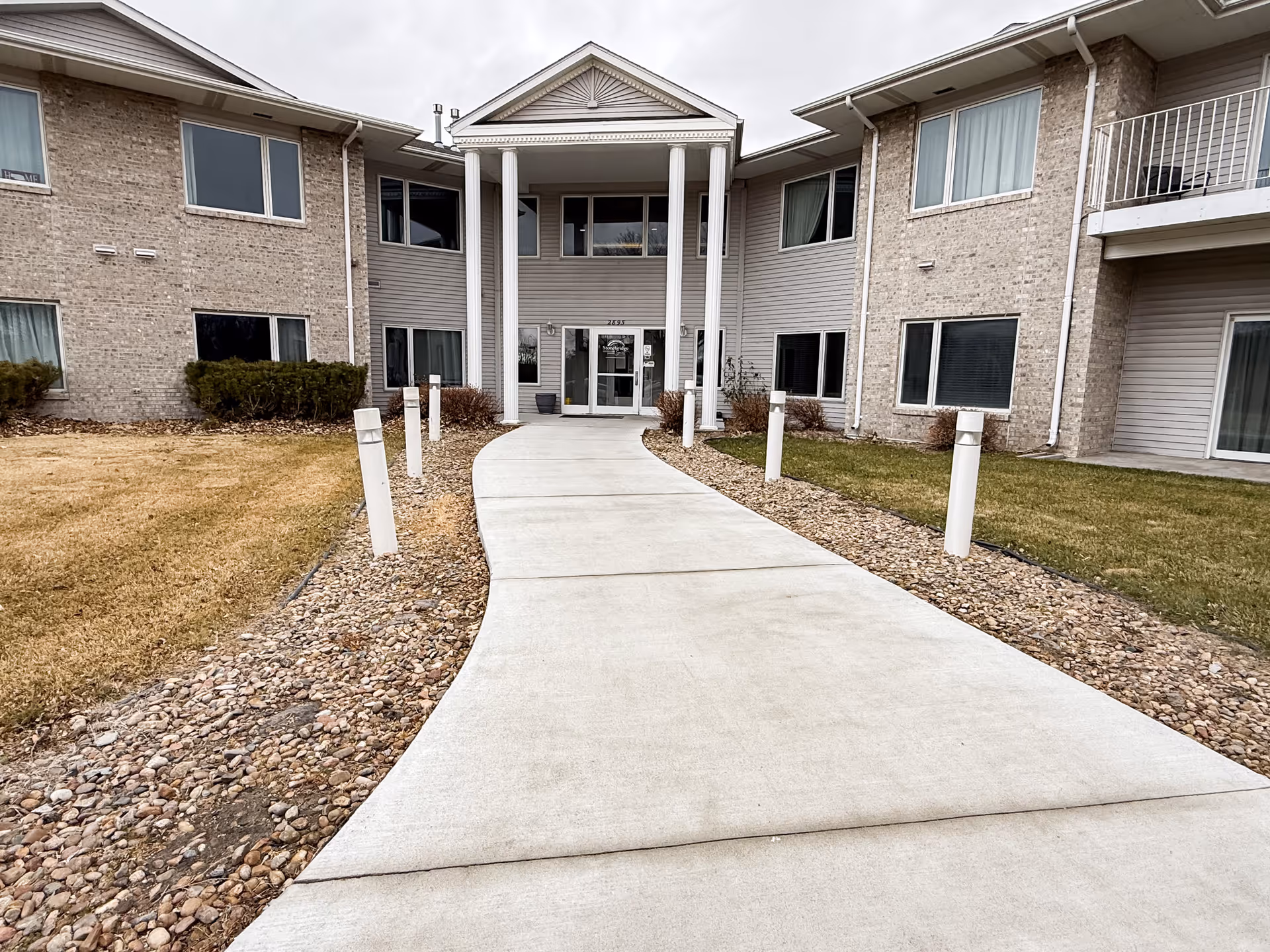 Curved concrete walkway to a senior living facility entrance with white pillars and brick facade