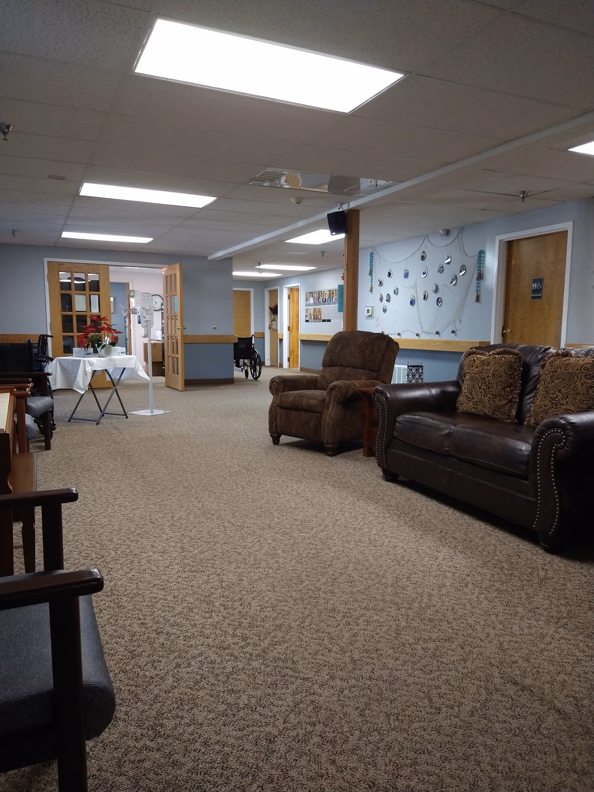 Cozy senior living lounge with brown leather sofas, chairs, and a small table near a hallway.