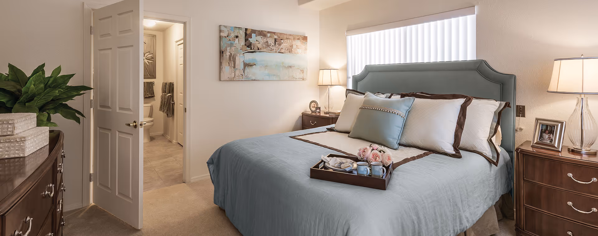 Well-lit bedroom with a blue upholstered headboard, a neatly made bed with pillows and a tray, bedside tables, and an open door leading to a bathroom.