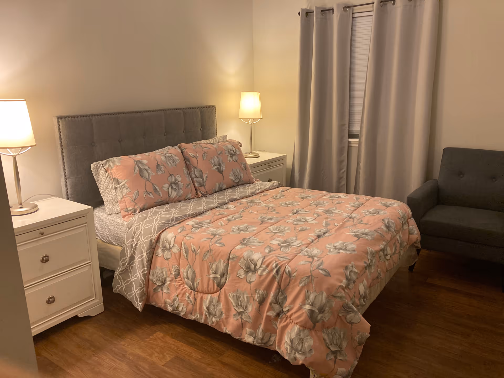 Cozy bedroom featuring a queen bed, two nightstands with lamps, curtains, and a grey sofa.