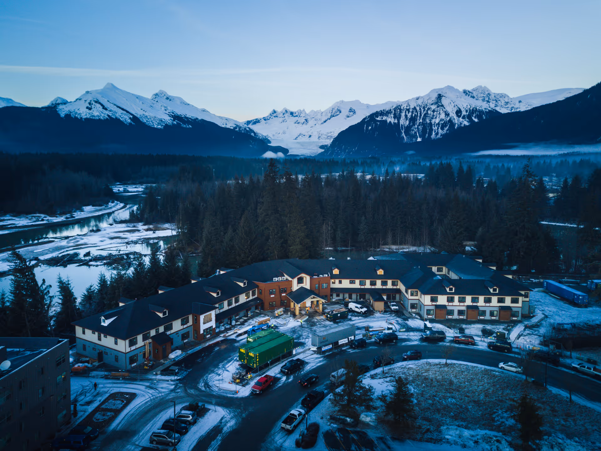 Aerial view of Riverview Senior Living facility surrounded by snow-covered ground and trees, with a backdrop of snow-capped mountains under a clear sky during dusk or dawn.