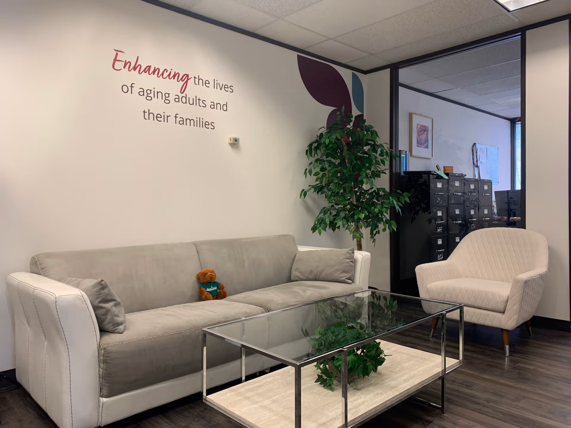 Waiting lounge with gray sofa, cream chair, glass coffee table, plants, and wall art.