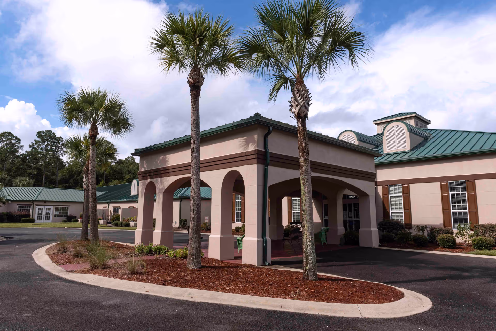 Exterior view of Magnolia Manor of St. Marys showing a covered entrance with arches, palm trees, and a building with green metal roofing and beige walls with brown shutters under a partly cloudy sky.