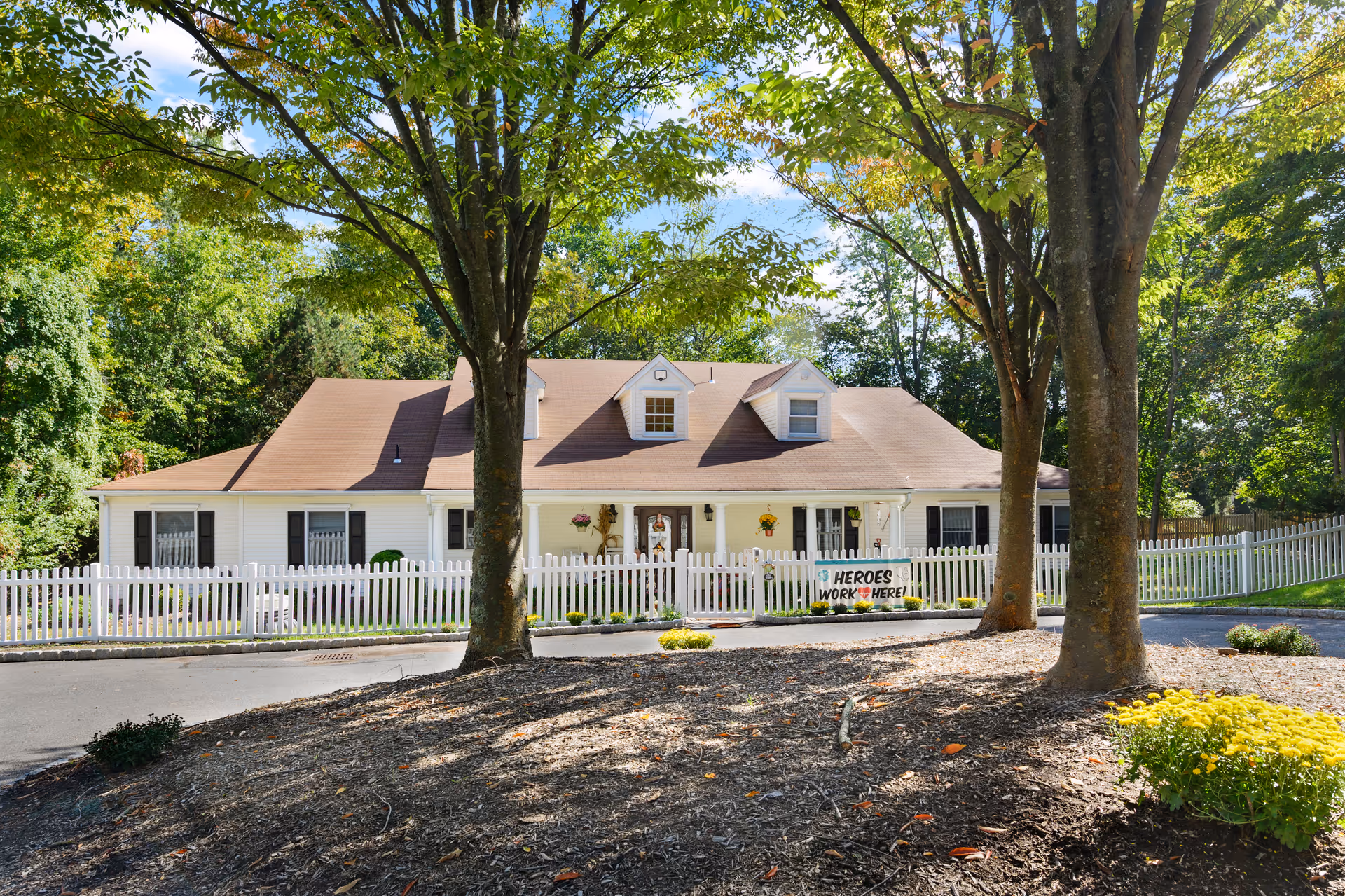 Front view of a single-story white house with a white picket fence, trees in the foreground, and a 'Heroes Work Here!' sign.