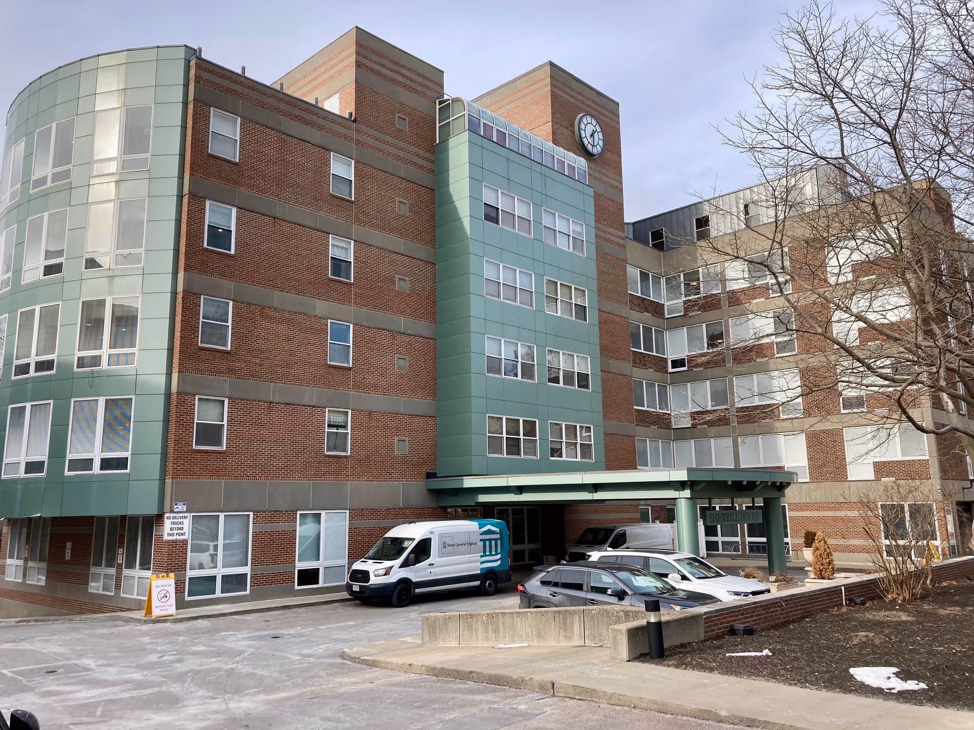 Exterior view of a multi-story brick and green-paneled senior living building with an entrance canopy, parked vehicles, and a clock tower.