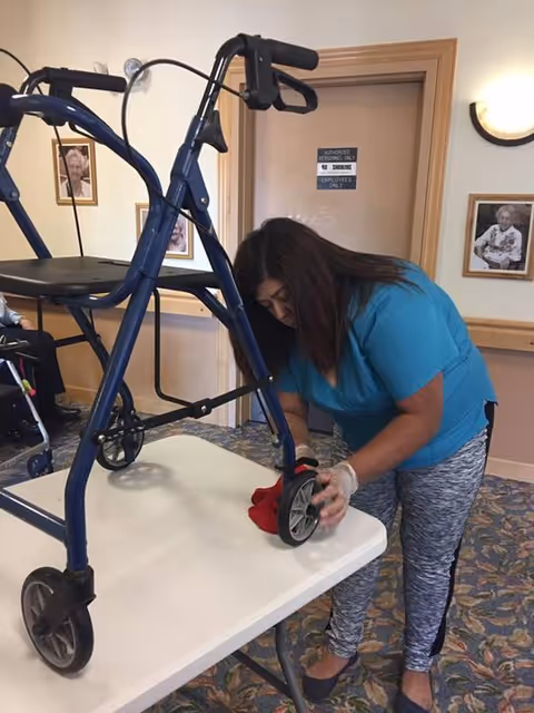 Caregiver in blue scrubs adjusts a walker on a table in a senior facility hallway.