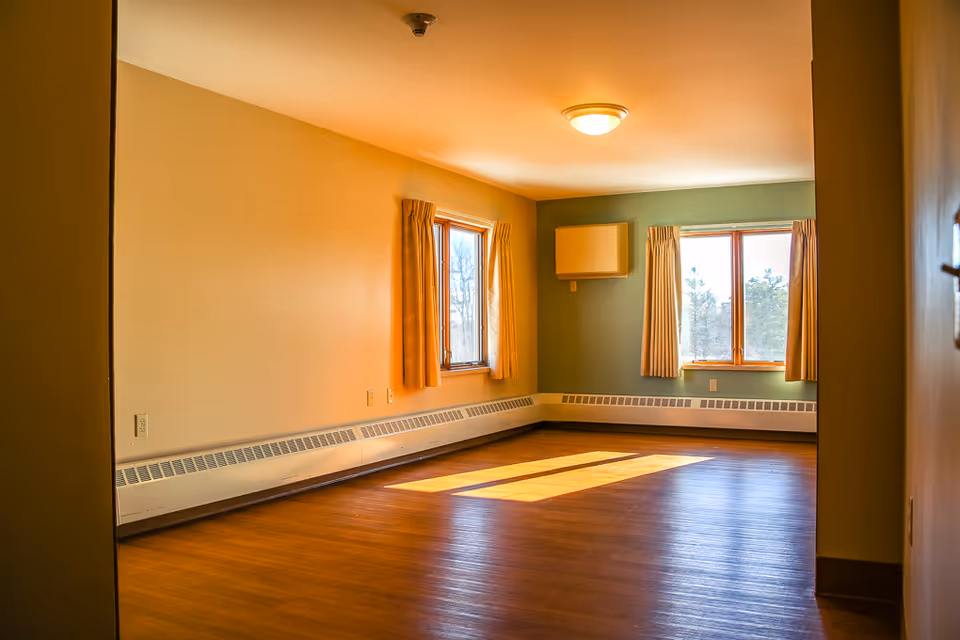 Empty senior living room with large windows, curtains, hardwood floor, and baseboard heaters.