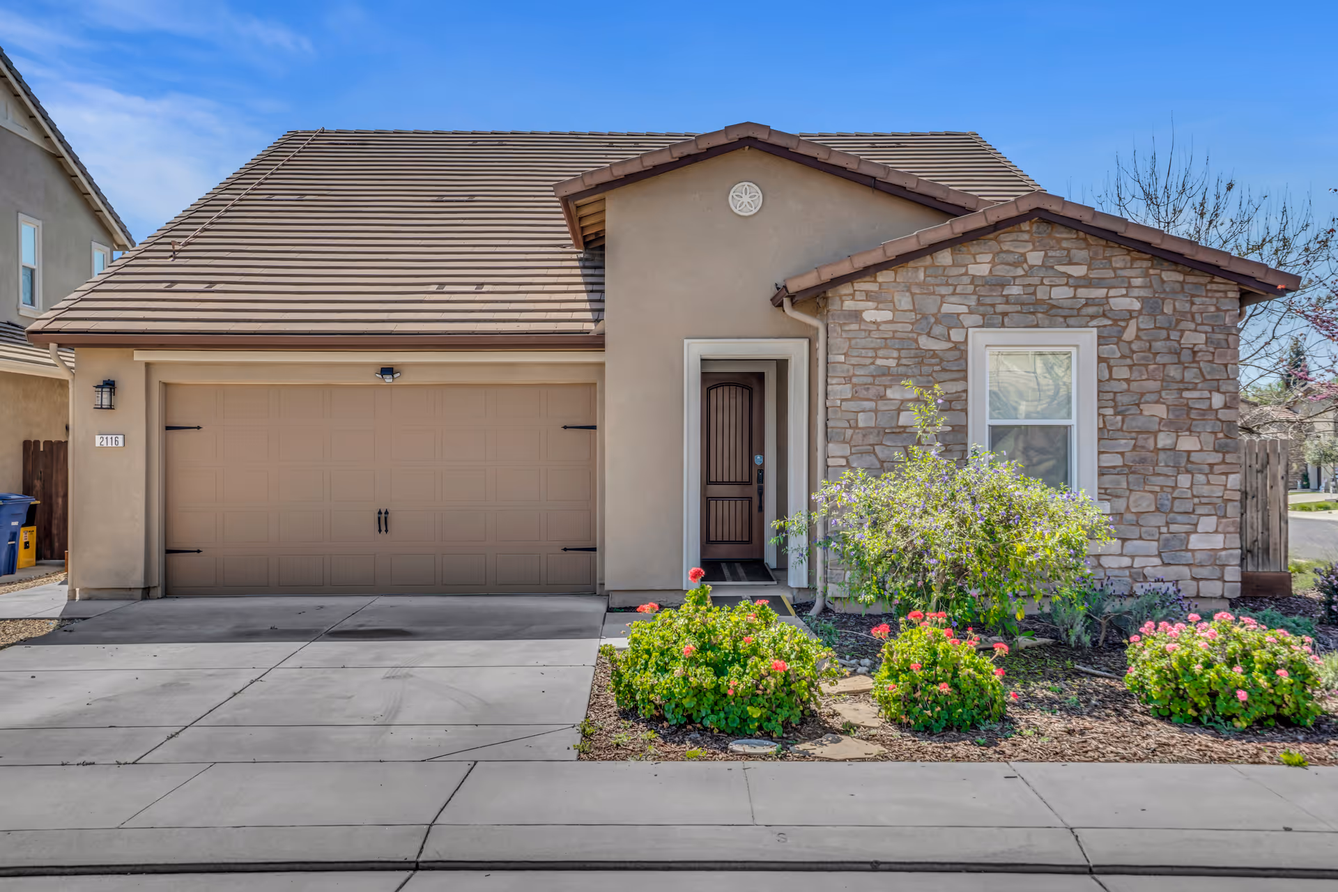 Front view of a beige stucco house with a stone accent wall, garage, and flowering shrubs.