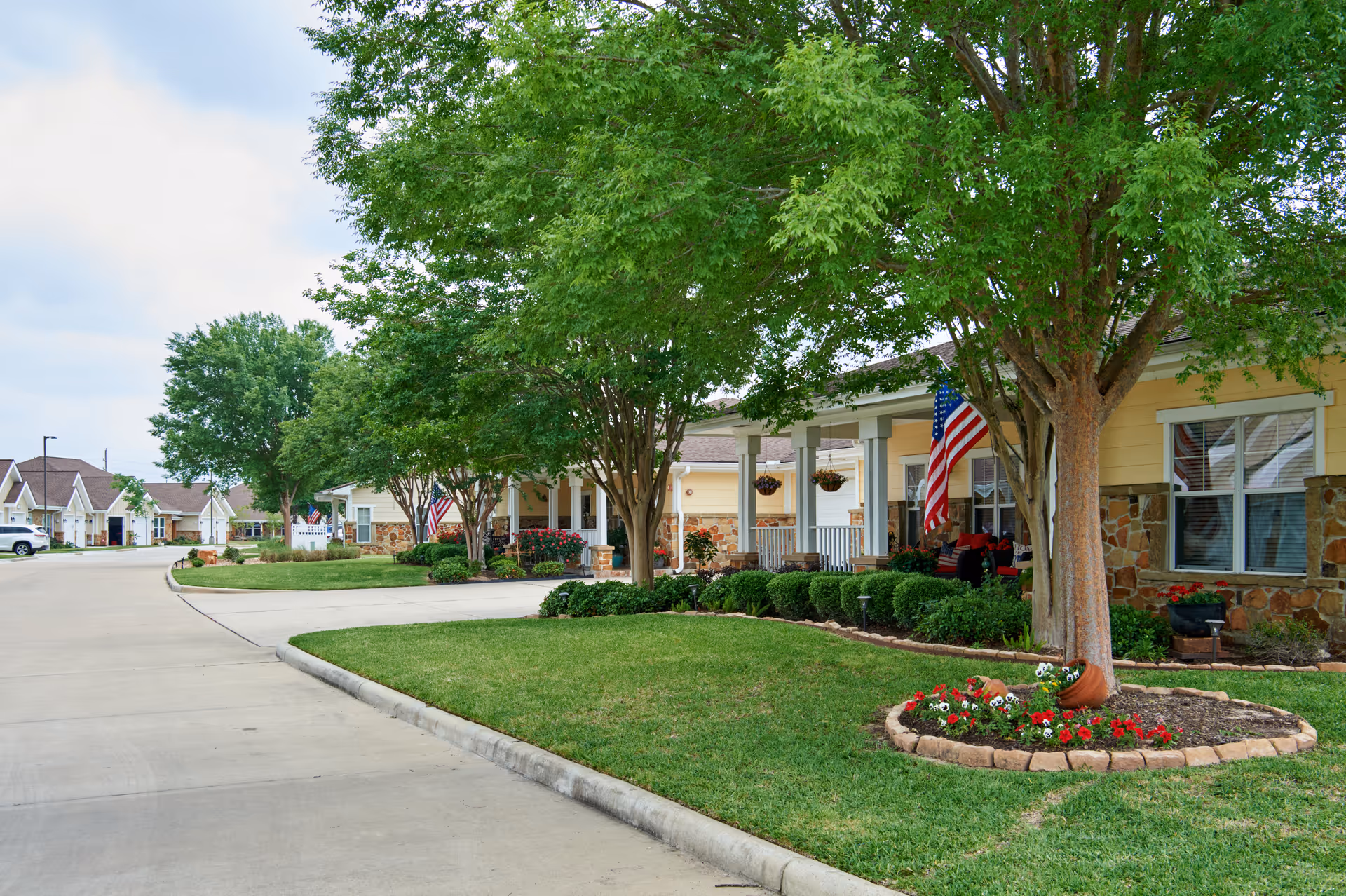 Exterior view of a senior living community with manicured lawn, trees, and flag-decorated porch.