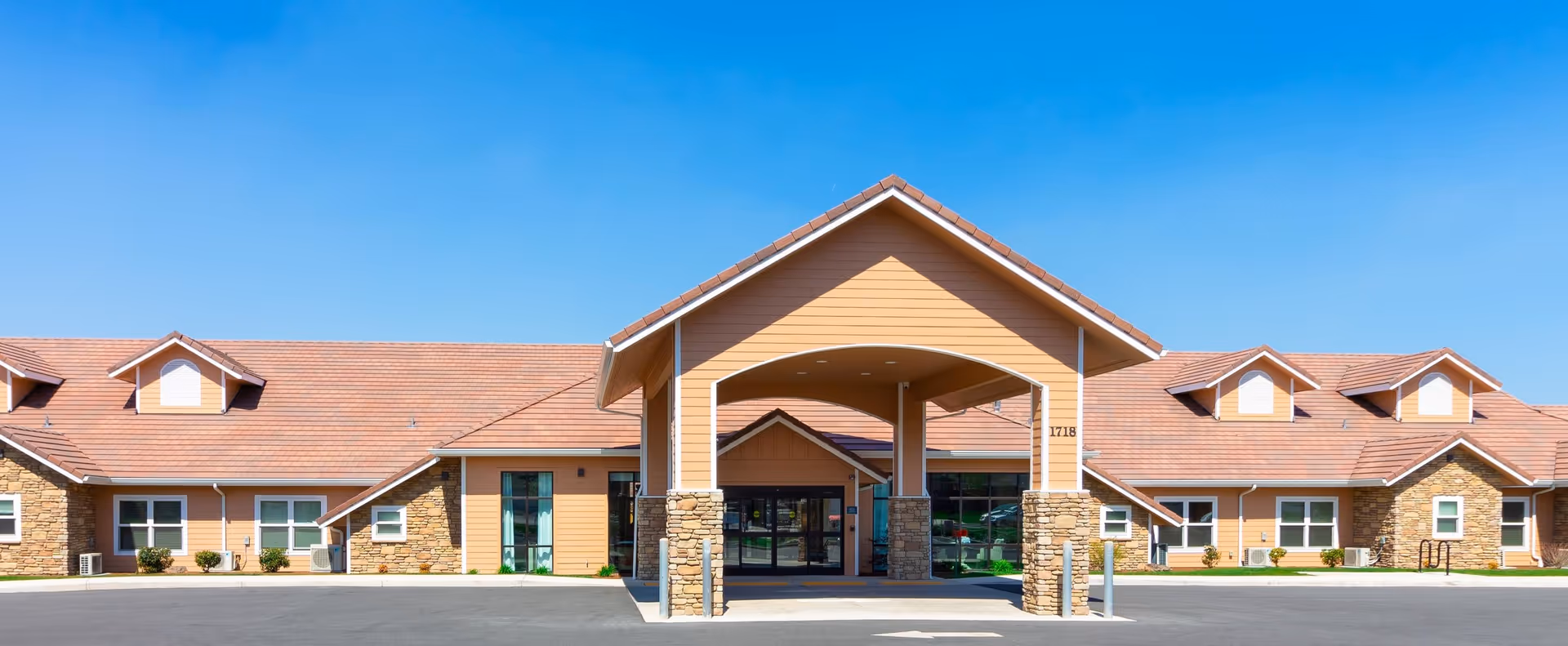 Front view of a single-story senior living facility with a covered porte-cochère entrance, stone accents, and a clear blue sky.