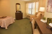 A cozy bedroom with a single bed covered in a patterned bedspread, a dark wooden dresser with a mirror, a beige armchair next to a window with blinds, a desk with a lamp, books, and a small plant, and a framed artwork on the wall.