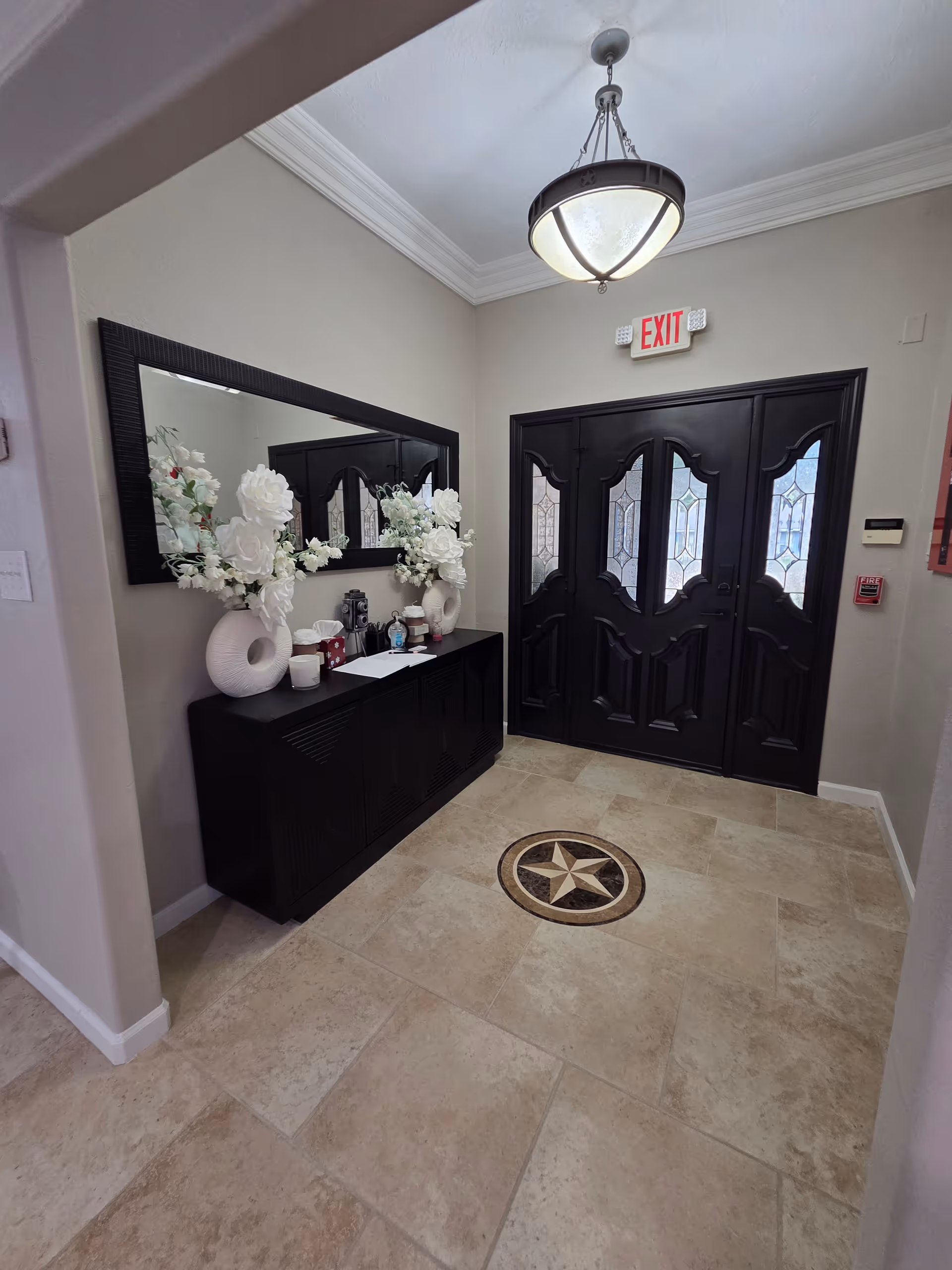 Indoor foyer with black console, large mirror, white flower arrangements, chandelier, and double doors.