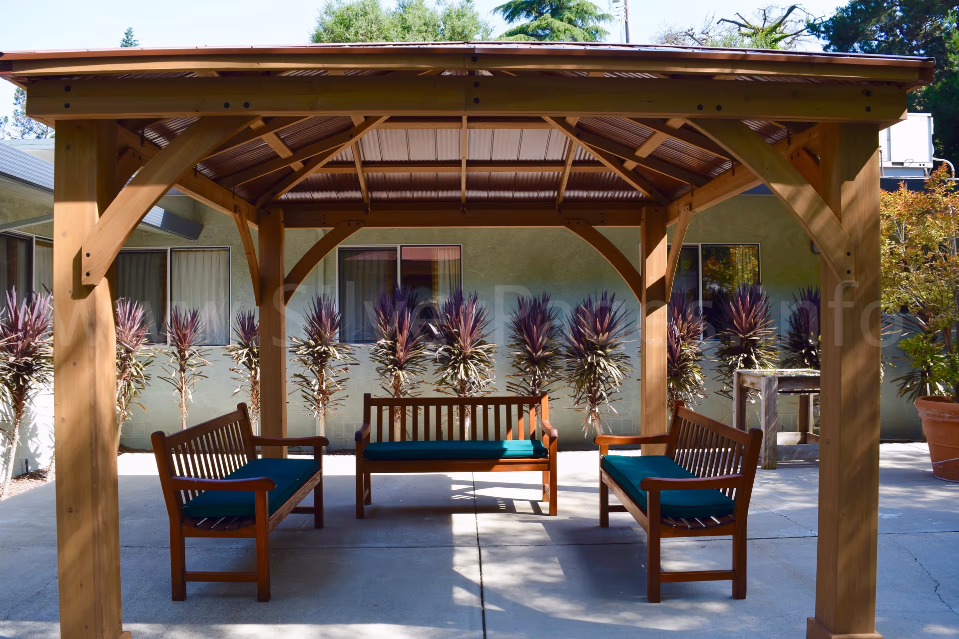 Wooden gazebo with benches and turquoise cushions in a sunny outdoor courtyard.