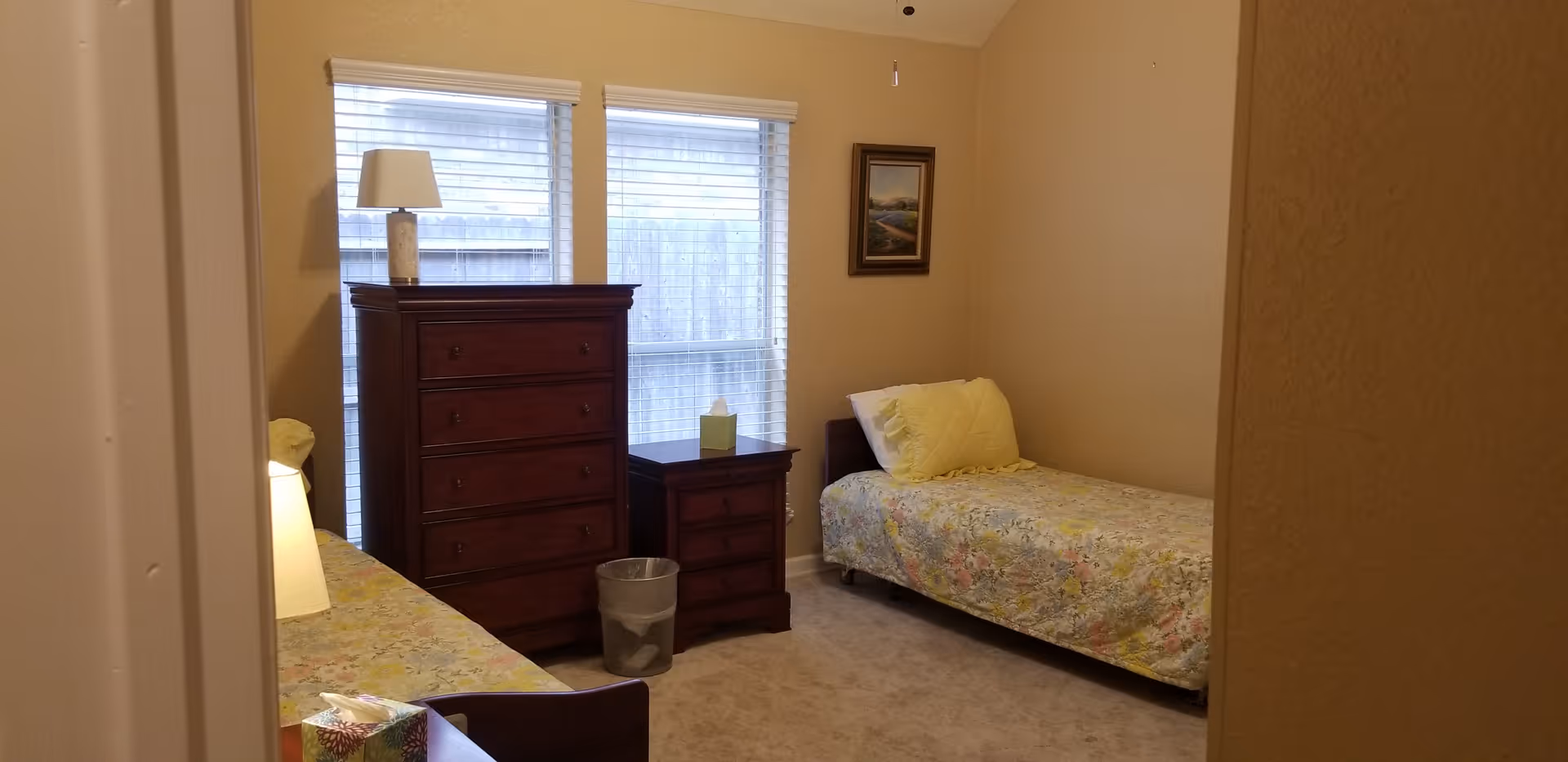 A small bedroom with two single beds covered in floral bedspreads, a wooden dresser and nightstand between two windows with blinds, a lamp on the dresser, a trash bin on the floor, and a framed picture on the wall.