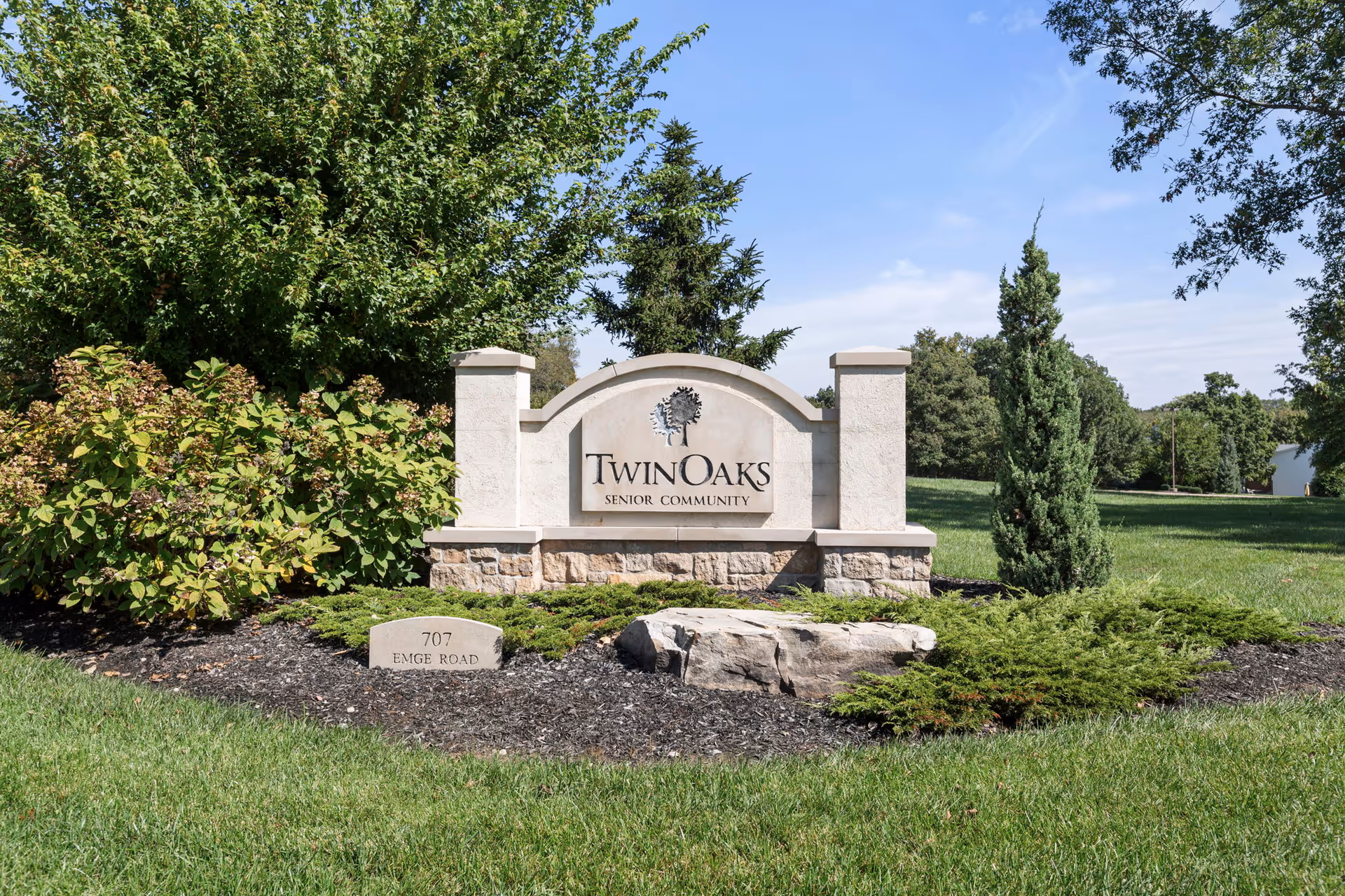 Outdoor view of a stone sign for Twin Oaks Senior Community surrounded by green grass, bushes, and trees under a clear blue sky.