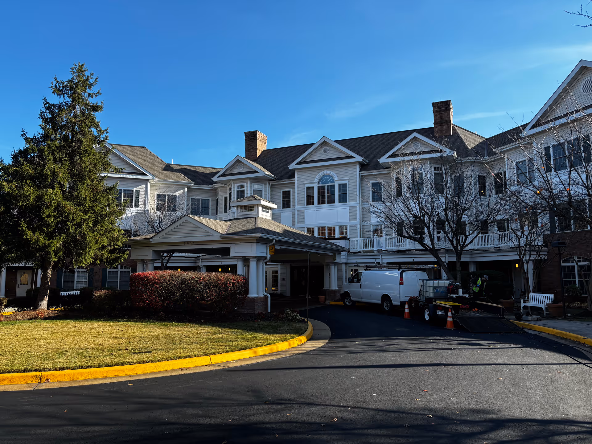 Three-story white senior living building with a covered entrance, driveway, and a parked white van under a clear blue sky.