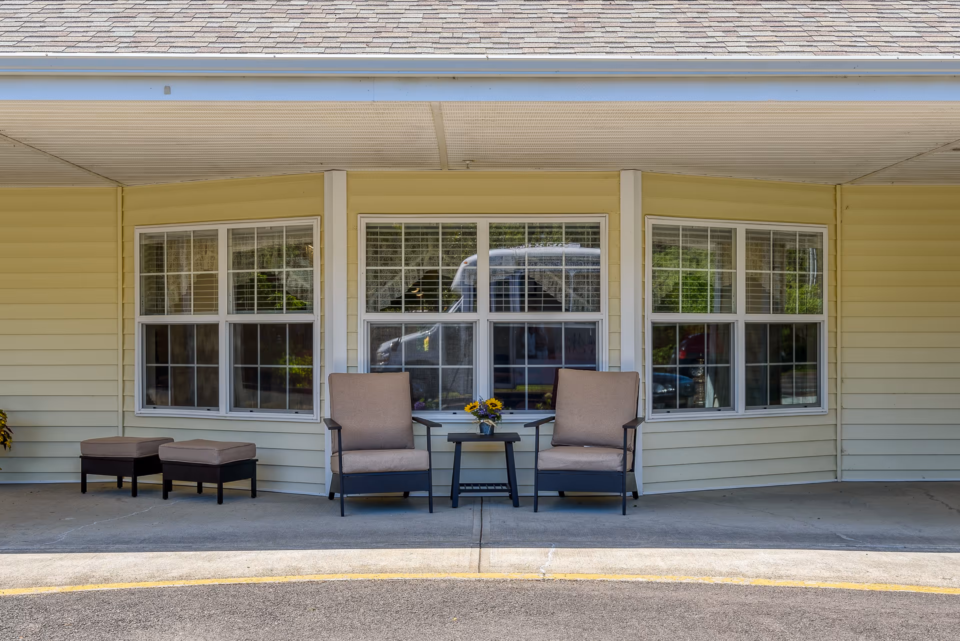 Covered building front with two cushioned armchairs, a small table with flowers, and three large windows.