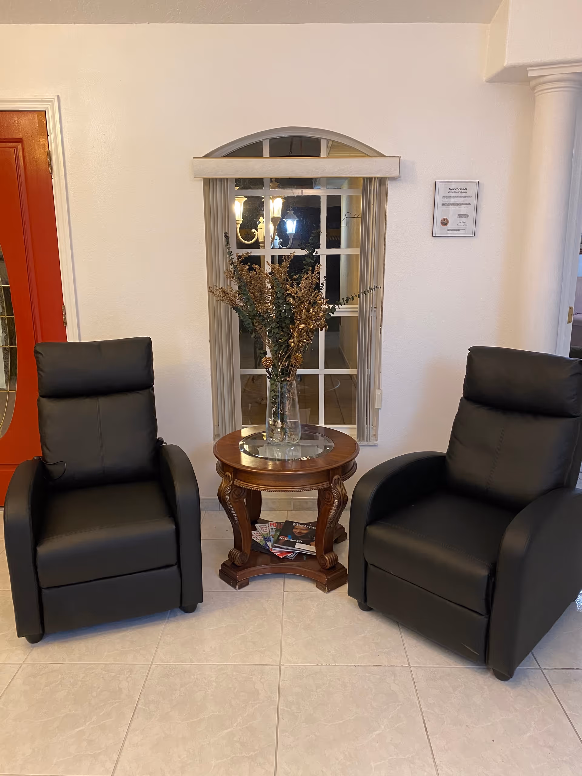 Two black leather recliner chairs positioned on either side of a round wooden side table with a glass top, which holds a clear vase with dried flowers. Behind the table is a window with vertical blinds and a decorative arch above it. A red door is visible on the left side of the image, and a framed document hangs on the wall to the right of the window.