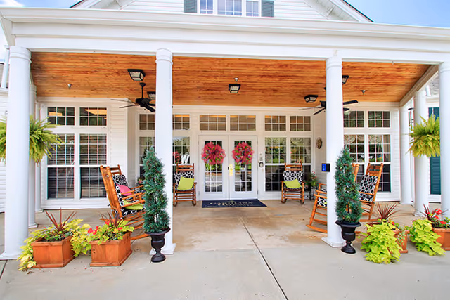 Entrance porch with white columns, rocking chairs, potted plants, and double glass doors adorned with wreaths.