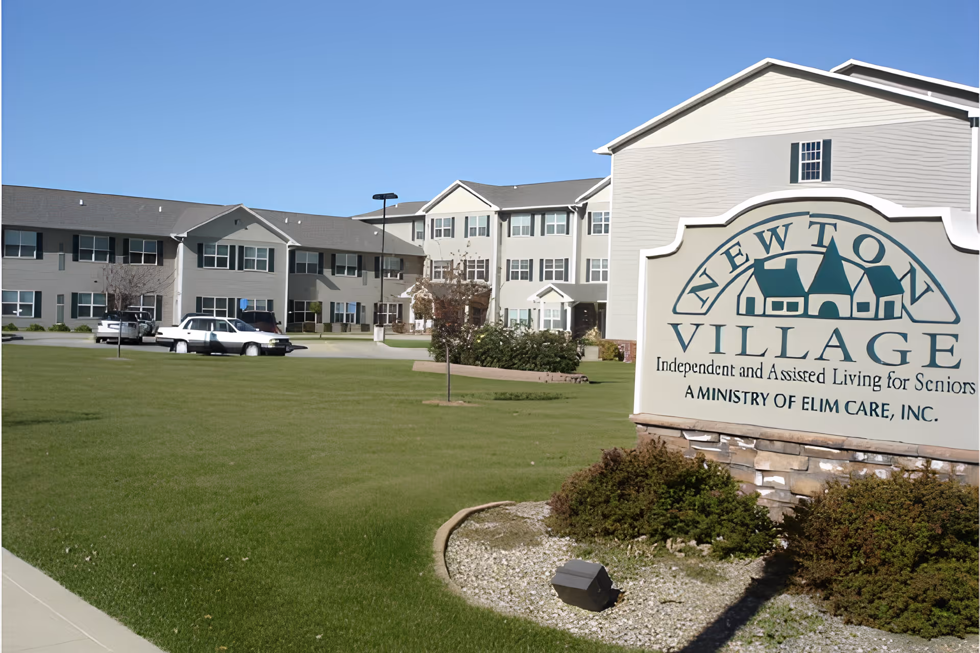 Exterior view of Newton Village, an independent and assisted living facility for seniors, showing a large green lawn, a sign with the facility's name, and a multi-story building with multiple windows and parked cars.