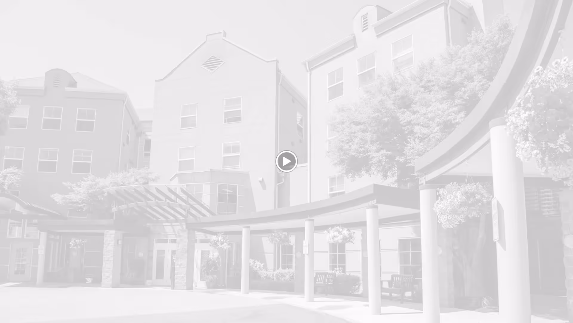 Exterior view of Chateau Pacific Retirement facility showing a multi-story building with multiple windows, a covered entrance with columns, hanging flower baskets, and benches outside.