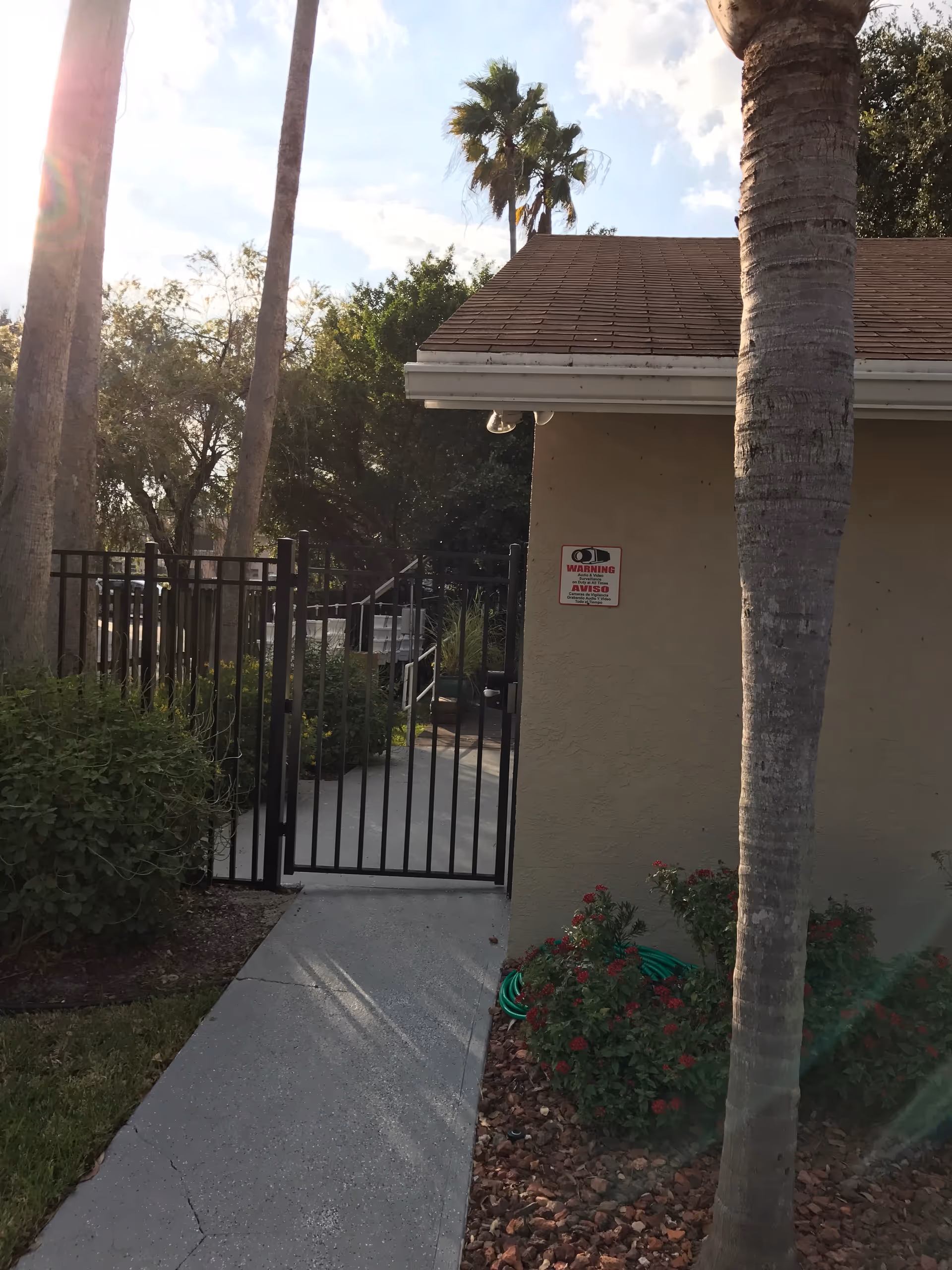 Exterior gate and walkway to a senior facility, with palm trees and landscaped shrubs.