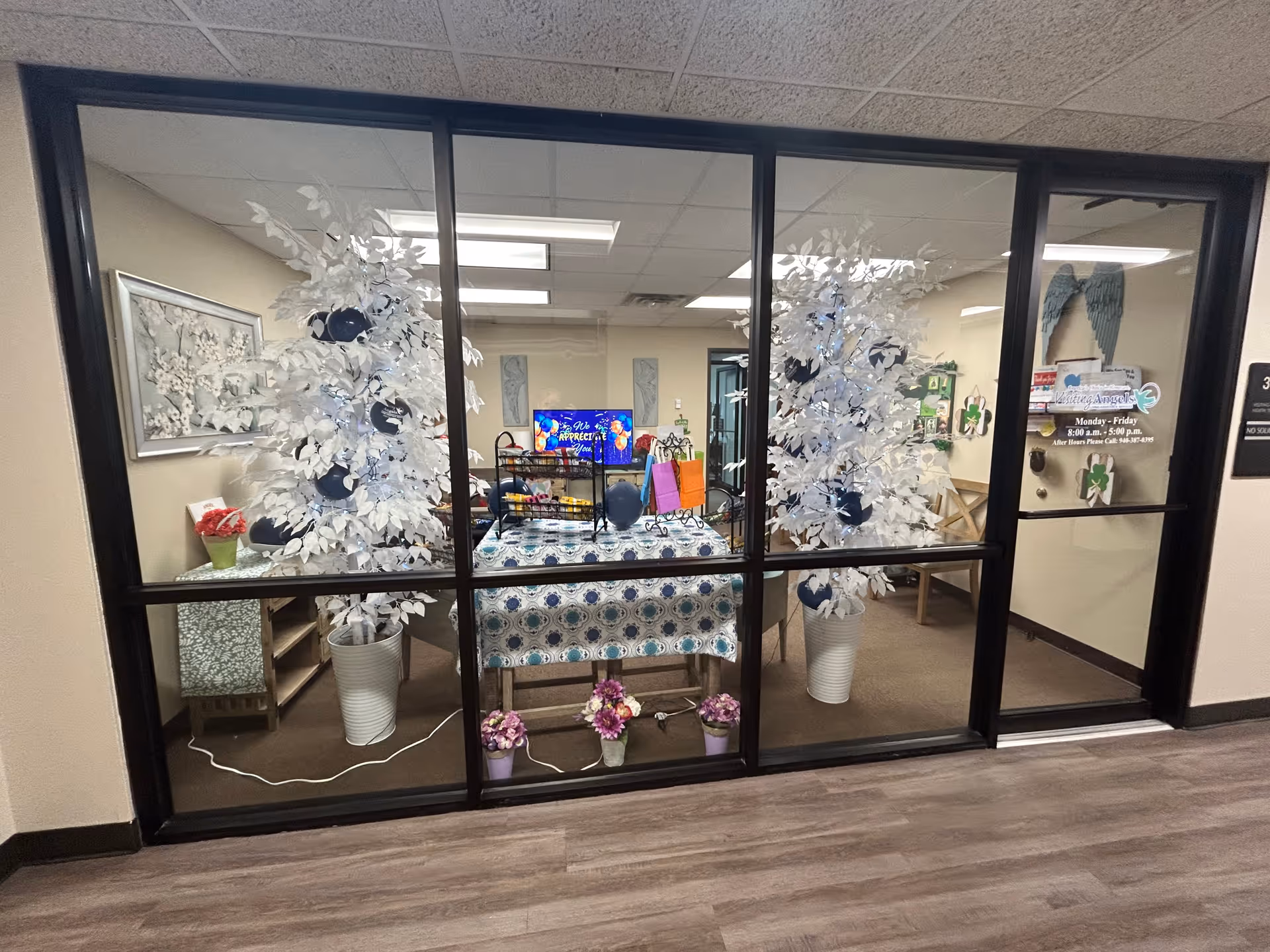 Inside a glass-walled common area with a decorated table and white artificial trees.
