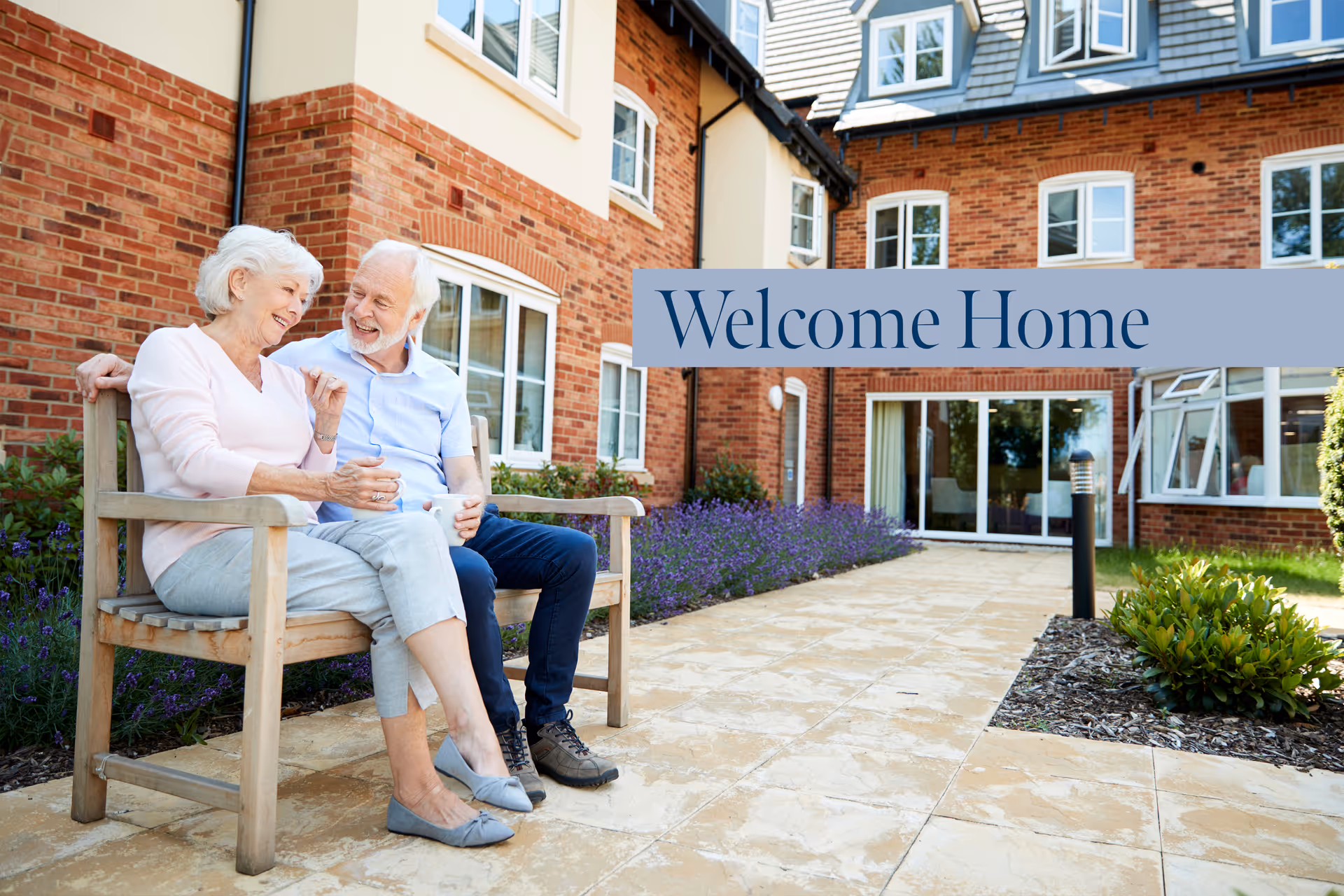An elderly couple sits on a bench outside a brick senior living building with a 'Welcome Home' banner.