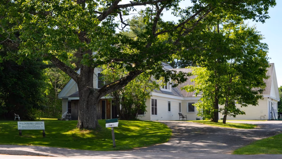 White senior living facility exterior with curved driveway, large trees, and a green lawn.