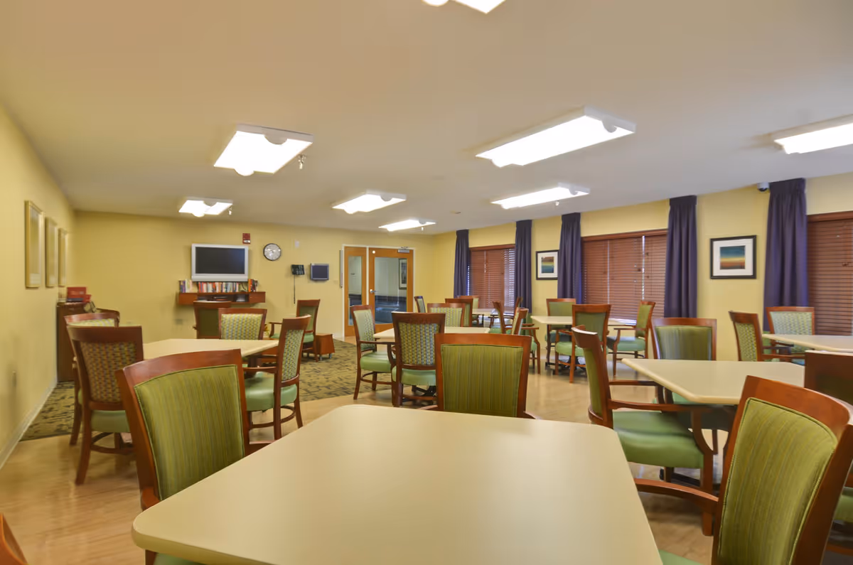 A well-lit dining room with multiple tables and chairs arranged neatly. The chairs have wooden frames with green upholstery. The room has yellow walls, several framed pictures, and windows with purple curtains and wooden blinds. A television and a clock are mounted on the far wall above a small bookshelf.