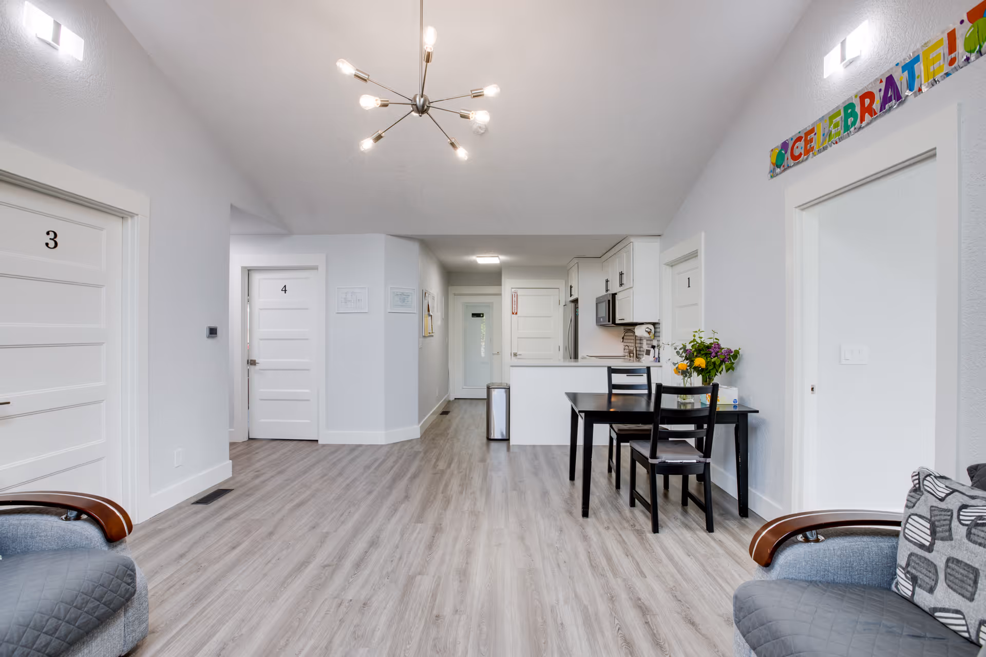 Bright communal interior with a small dining table and kitchenette, numbered resident doors, and seating.