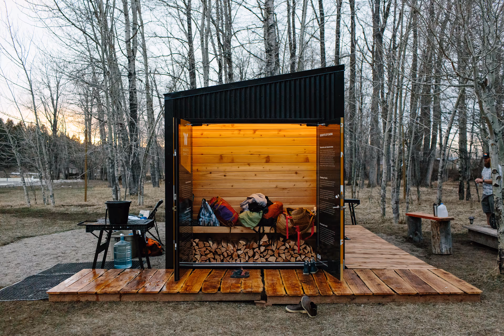 Small black sauna shed on a wooden deck in a forest with open doors.