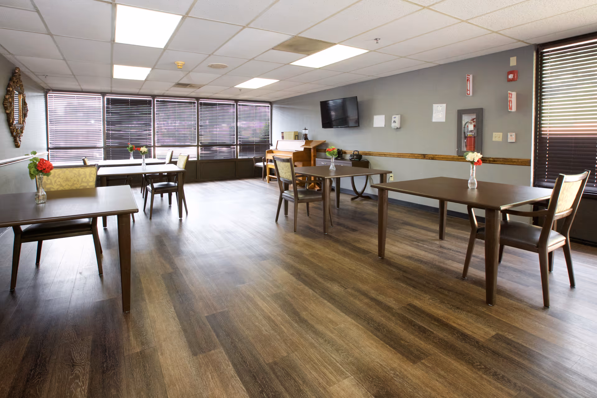 A bright dining room with several wooden tables and chairs spaced apart. Each table has a small vase with flowers. Large windows with blinds cover one wall, and a flat-screen TV is mounted on another wall. The room has wood flooring and a ceiling with recessed lighting.