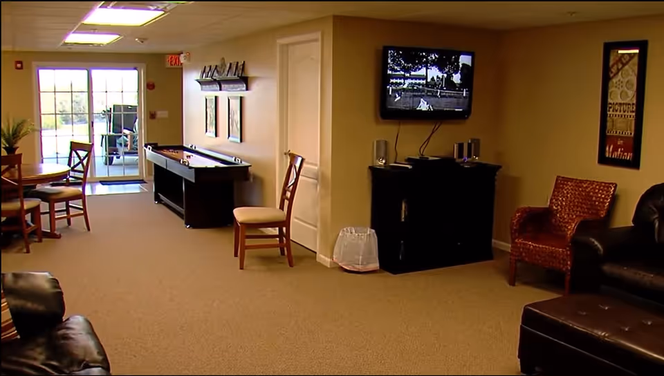 A common area in Ambrose Park Residential Care Facility featuring a carpeted floor, a round wooden table with chairs near a glass door leading outside, a shuffleboard table against the wall, a wall-mounted TV above a cabinet with speakers, and several chairs including a leather sofa and a patterned armchair.