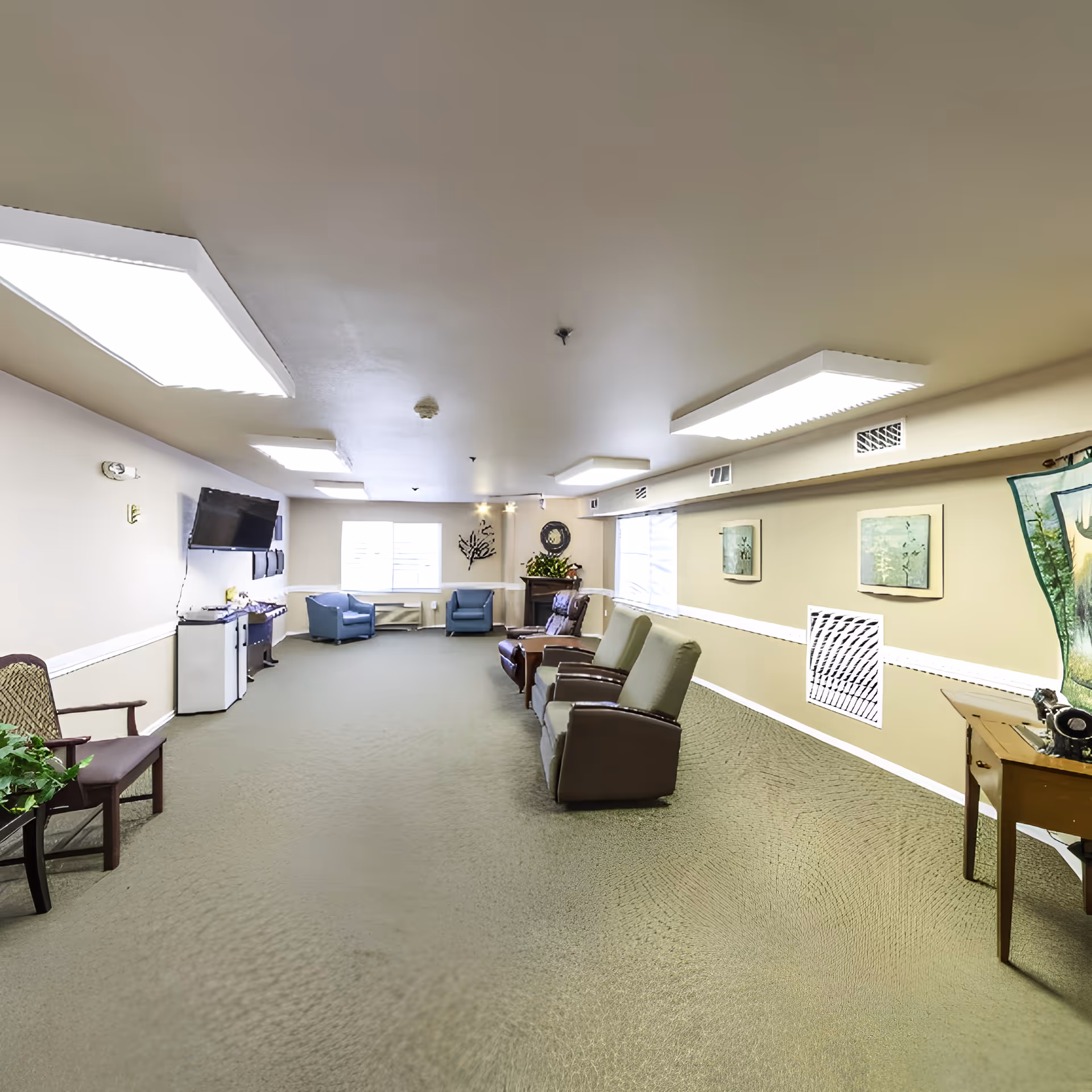 A spacious common area with beige walls and carpeted floor, featuring several armchairs and recliners arranged along the walls. There is a flat-screen TV mounted on the left wall above a small cabinet and a mini fridge. The room is well-lit with large rectangular ceiling lights and has windows at the far end letting in natural light. Decorative artwork and a plant are visible on the right wall and near the windows.