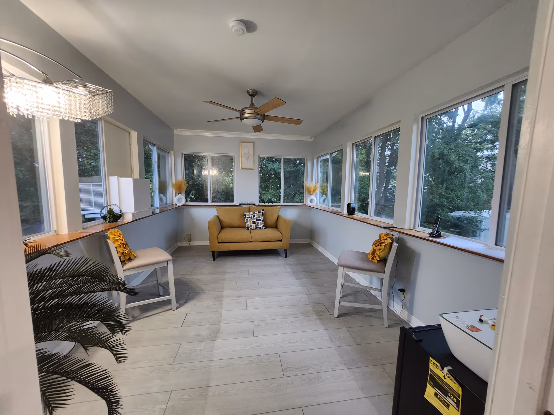 A bright sunroom with large windows on three sides showing green trees outside. Inside, there is a mustard yellow loveseat with a patterned cushion in the center against the far wall. Two beige chairs with yellow decorative pillows are placed on either side of the room. A ceiling fan with wooden blades is mounted on the white ceiling. The floor is light-colored wood or wood-like tile. A small black refrigerator with a white bowl on top is visible in the foreground on the right. A leafy green plant is partially visible on the left side near the entrance.