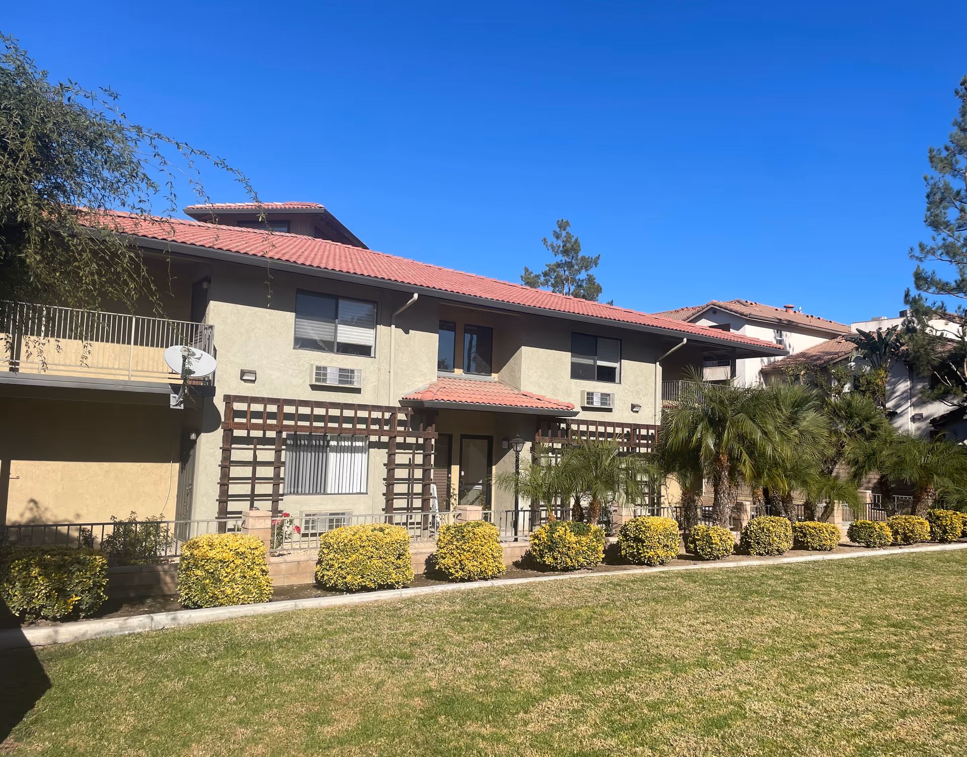 Two-story residential building with a red tile roof, balconies, palm trees and a manicured lawn in front.