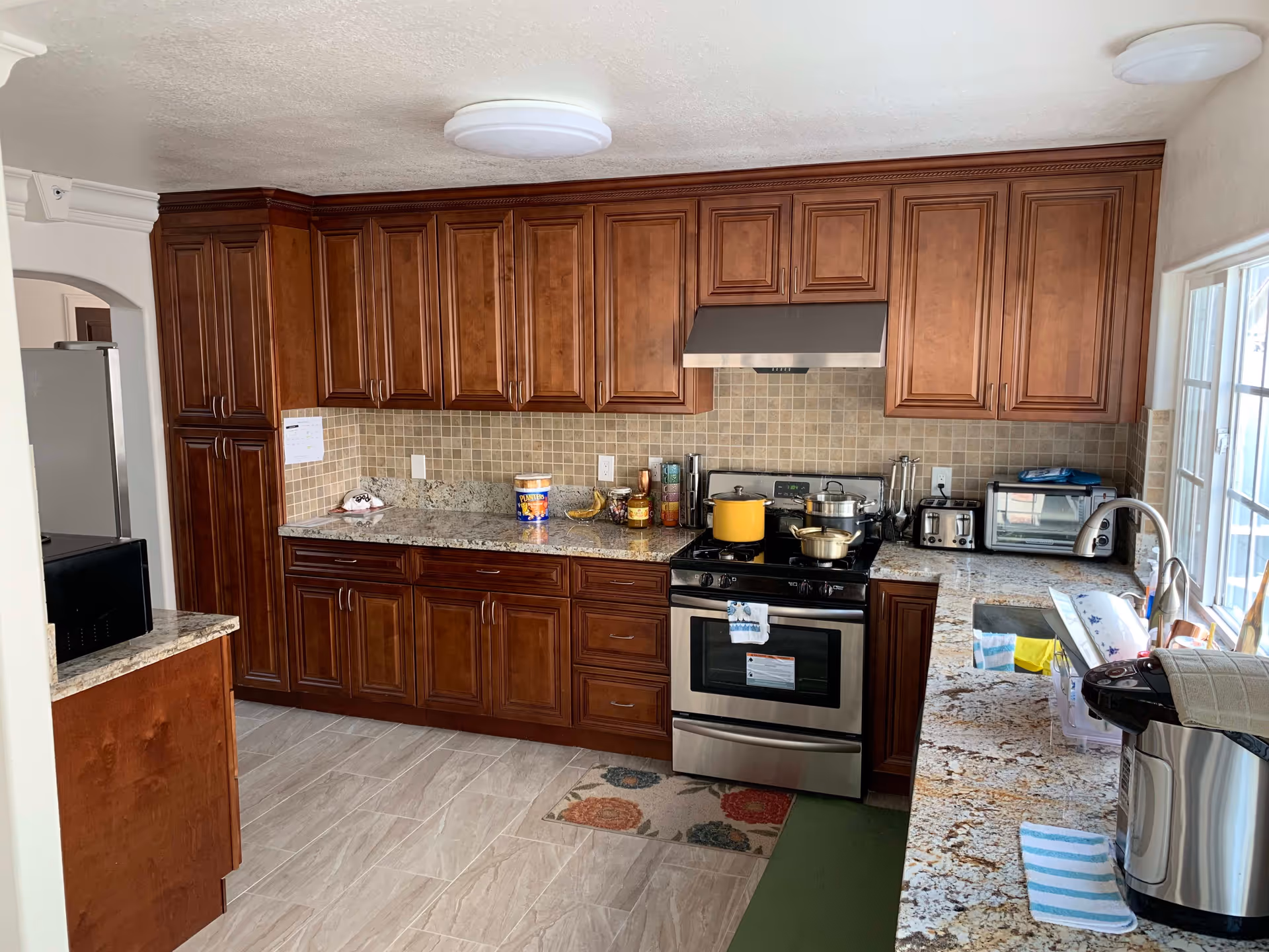 A kitchen with wooden cabinets, granite countertops, and tiled backsplash. The kitchen features a stainless steel stove with pots on it, a toaster, a microwave, a rice cooker, and a window letting in natural light. The floor is tiled and there are small rugs near the stove and sink area.