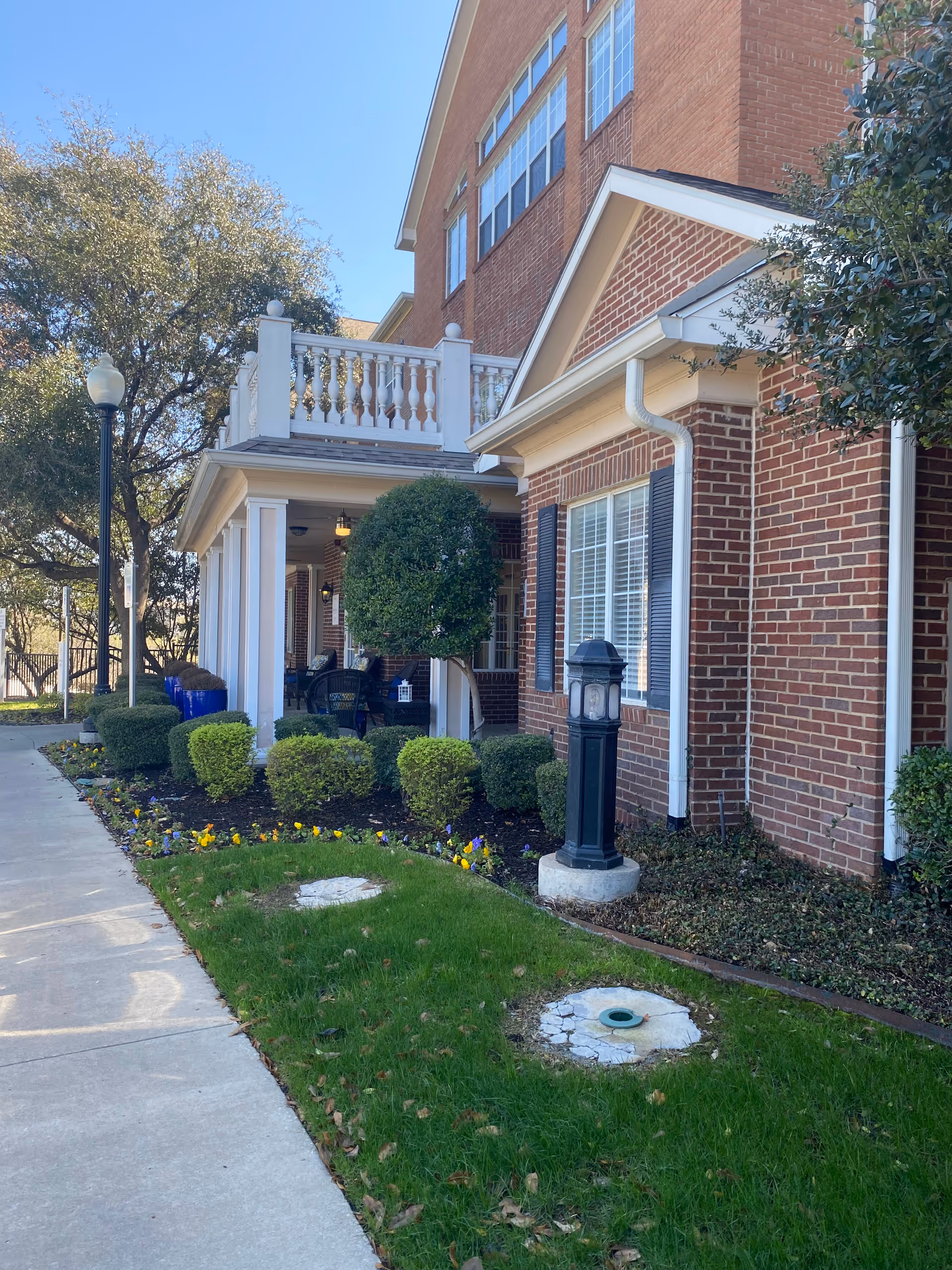 Brick senior living building exterior with white porch, columns, balcony, and neatly landscaped front yard.