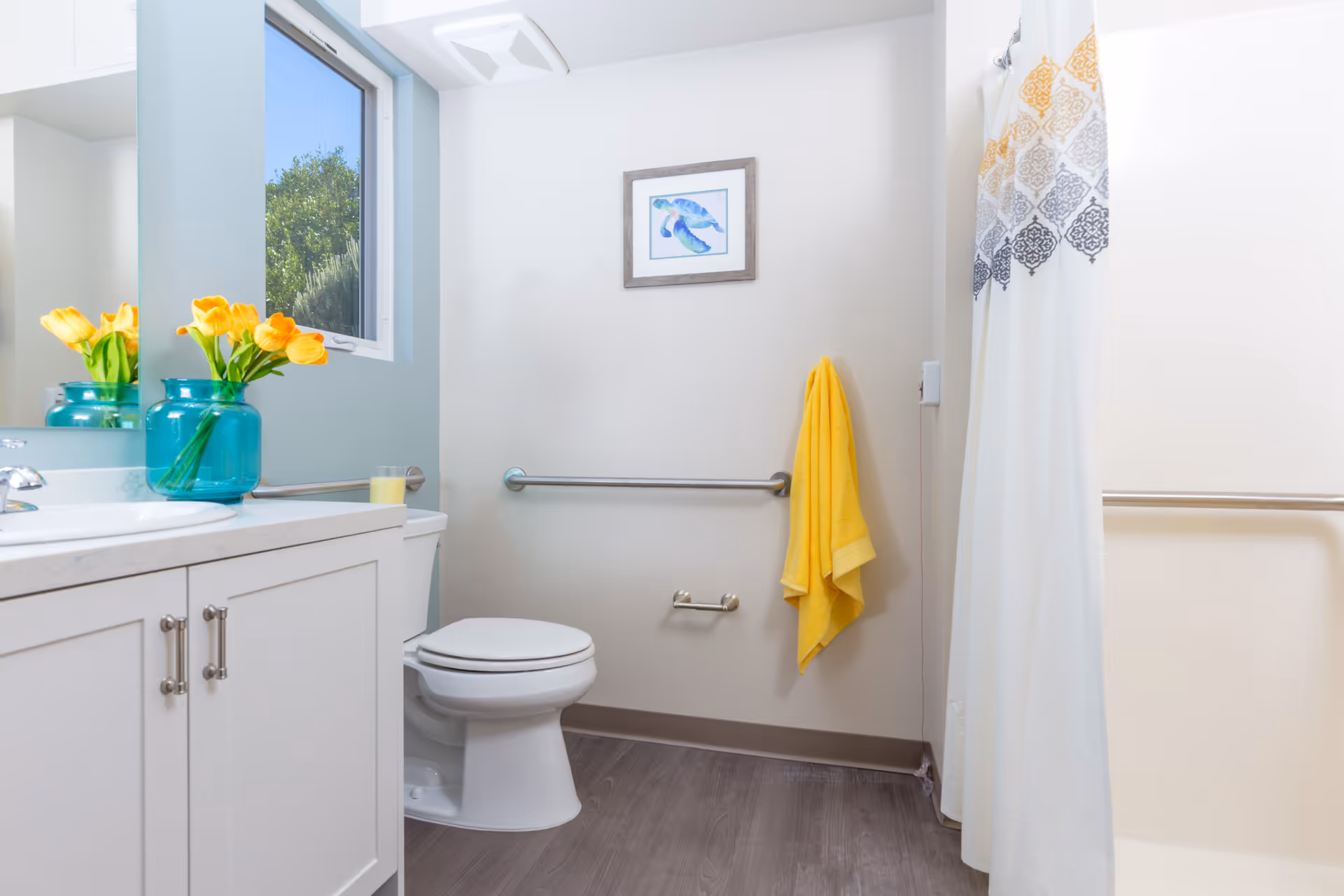 Bright accessible bathroom with a toilet, vanity topped with flowers, grab bars, and a shower with a patterned curtain.